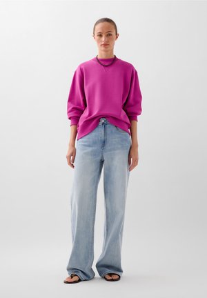 Magenta oversized sweatshirt en lichtblauwe wijde jeans. Getextureerde stof met geribde manchetten en een relaxte pasvorm. G gedragen met zwarte flip-flops.