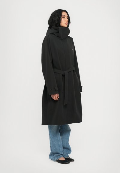 Black long coat with a high collar, belted waist, waterproof material, and side pockets. Worn over blue wide-leg jeans and black flats.