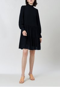 Musta pitkähihainen mekko, jossa on korkea kaula-aukko, kerroksellinen hame ja pehmeä kangas. Yhdistetty vaaleanruskeisiin avokkaisiin. Yksinkertainen, elegantti muotoilu.