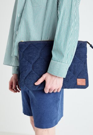 Personne portant une chemise rayée verte et blanche et un short bleu tenant une pochette matelassée bleu foncé avec une étiquette marron et une fermeture éclair.