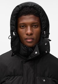 Fiatal férfi rövid göndör hajjal, fekete kapucnis téli dzsekit visel, amelyen fém patentgombok és húzózsinórok vannak, előre néz.