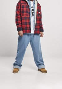 Rot und marineblau kariertes Jackett über einem weißen T-Shirt, kombiniert mit hellblauen lässigen Jeans und sandfarbenen Schuhen. Die Jacke hat ein Fleece-Futter und einen Reißverschluss.