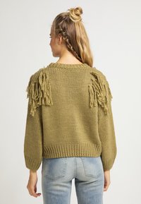 Sweter w oliwkowym kolorze z frędzlami na ramionach, okrągłym dekoltem i ściągaczem na dole. W zestawieniu z jasnoniebieskimi dżinsami, widok z tyłu.