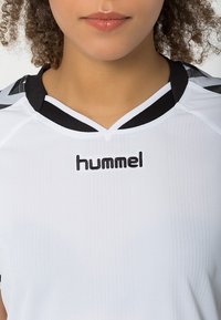 Camisa deportiva blanca con un cuello acanalado negro, que presenta el logo "hummel" en negro. Textura de líneas verticales en la parte frontal.