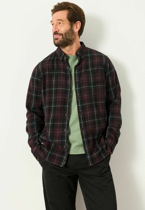 Mann mit Bart, der ein dunkles kariertes Hemd über einem grünen T-Shirt und schwarzen Hosen trägt, steht mit den Händen in den Taschen vor einer weißen Wand.