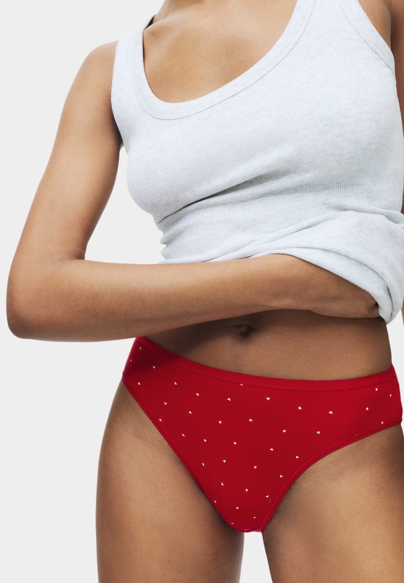 Femme portant des sous-vêtements rouges ornés de petits cœurs blancs et un débardeur blanc côtelé, ajustant le haut en le soulevant légèrement.