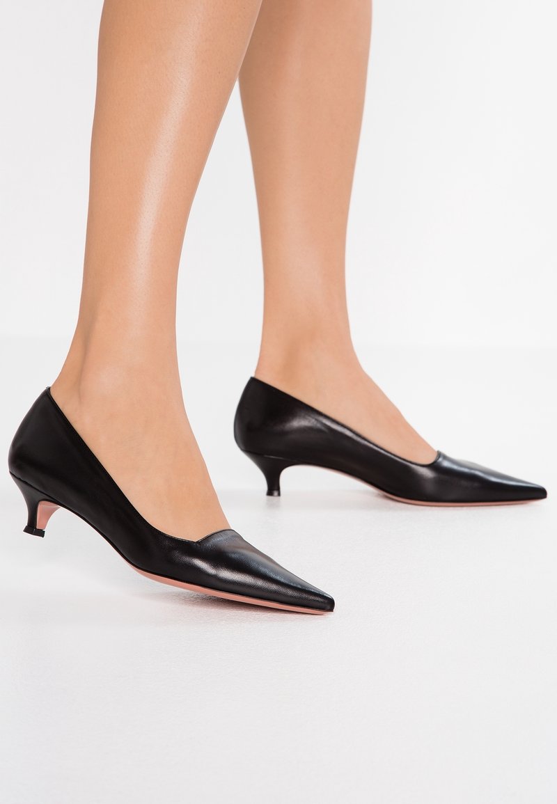 Femme portant des chaussures en cuir noir à talons kitten pointues, debout sur une surface blanche.