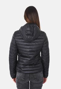 Donna con lunghi capelli castani, che indossa un giubbotto a cappuccio nero imbottito e jeans grigio scuro, vista di spalle su uno sfondo bianco.