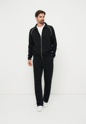 Férfi, fekete cipzáras dzsekit fehér csíkozással, fekete nadrágot, fehér pólót és fehér sportcipőt visel, fehér háttér előtt áll.