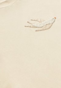 Beige katoenen T-shirt met een textuur, geborduurd vogelontwerp in het wit met een klein oranje detail op de snavel, gecentreerd op het borstgebied.