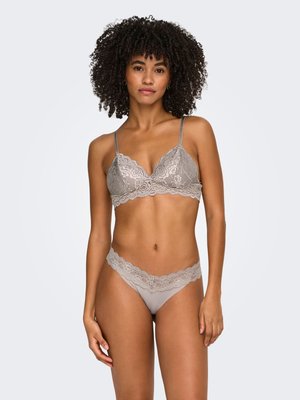 Woman with curly hair wearing grey lace bralette and matching lace-trimmed underwear, standing against a plain light background.