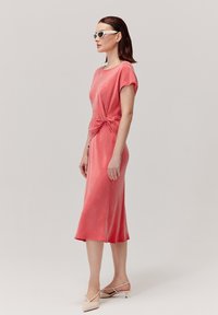 Sukienka do kolan w kolorze koralowym, z krótkimi rękawami, z węzłem w talii, wykonana z miękkiego materiału; modelka zestawiła ją z białymi okularami przeciwsłonecznymi i beżowymi obcasami.