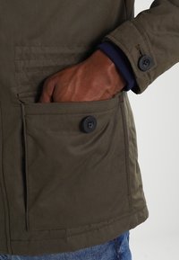 Veste vert olive avec une poche avant boutonnée, un tissu texturé et un poignet en tricot bleu marine visible au niveau du poignet.