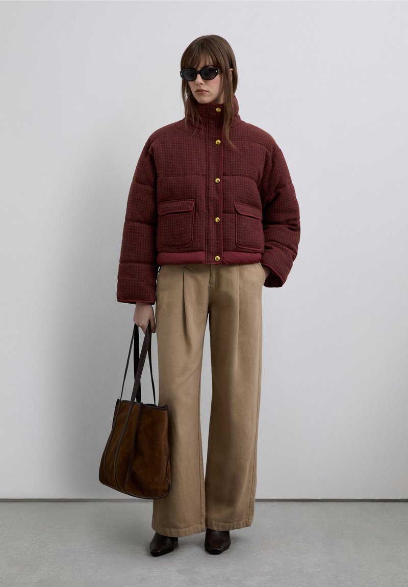 Veste matelassée bordeaux avec boutons dorés et deux poches avant, associée à un pantalon beige à jambes larges et un sac fourre-tout en daim marron.