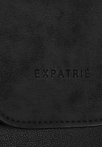 Černá kožená taška s texturovaným povrchem, s vyraženým logem "EXPATRIÉ" a hladkým lemováním.