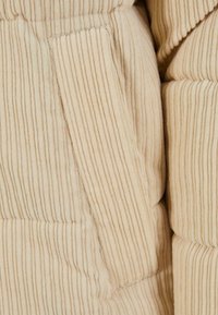 Béžová manšestrová látka s vertikálním žebrováním a detailem boční kapsy, součást nadýchaného svrchního oděvu.