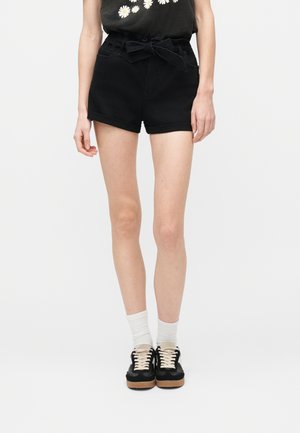 Osoba ubrana w czarne, wysokie spodenki z paskiem zawiązanym na kokardę, białe skarpetki oraz czarne trampki z białymi sznurowadłami, stojąca na tle jednorodnego tła.