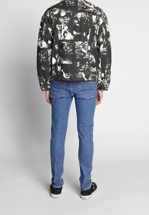 Muž v černo-bílé džínové bundě s potiskem, modrých úzkých džínách a černých teniskách, stojící zády k divákovi na jednobarevném pozadí.