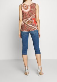 Haut sans manches rouge et rose à motif, associé à un pantalon capri en denim foncé et des sandales à talons argentées. Motif paisley remarquable sur le haut.