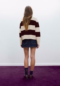 Pleteni pulover s kremnimi in maroon črtami, v kombinaciji s temno modrim krilom. Model nosi črne nogavice in čevlje, obrnjena je stran od gledalca.