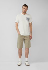 Witte katoenen t-shirt met een blauwe graphic op de borst, gecombineerd met beige denim shorts. Model draagt witte sokken en sneakers met zwarte accenten.