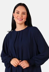 Blouse bleu marine foncé avec un devant plissé, un col rond et des poignets smockés. Tissu doux, coupe ample, avec des détails texturés.