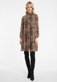 Frau in einem langärmeligen, knielangen Kleid mit Leopardenmuster, Rüschensaum am Hals und schwarzen hochgekrempelten Stiefeln, die vor einem weißen Hintergrund steht.