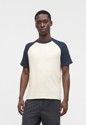 Uomo che indossa una t-shirt bianca a maniche corte con maniche raglan blu navy e pantaloni grigio scuro, in piedi davanti a uno sfondo chiaro uniforme.