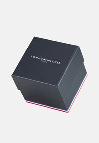 Caja de reloj negra con tapa ligeramente elevada, que presenta el logotipo "TOMMY HILFIGER" y acentos en rojo, blanco y azul en los bordes de la base.