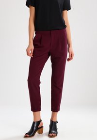 Pantalons bourgogne ajustés avec pinces, jambe fuselée et longueur cheville. Associés à un top noir à manches courtes et des talons block ouverts noirs.