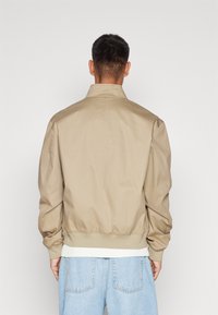 Bež bomber jakna z rebrastimi manšetami in pasom. Narejena iz gladkega materiala, ima klasičen ovratnik in sproščen kroj, oblečena čez svetlo denim.