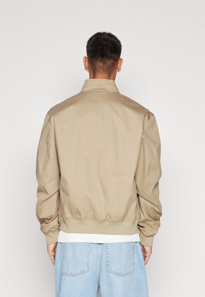 Veste bomber beige avec des poignets et une ceinture côtelés. Fabriquée en tissu lisse, elle présente un col classique et une coupe décontractée, portée sur un denim clair.