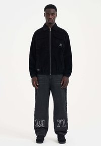 Mustapaneelinen corduroy-takki etutaskuilla, vetoketjukiinnityksellä ja joustavalla helmalla. Tummansiniset farkut sivukaitaleilla ja brodeeratulla "1971"-yksityiskohdalla.