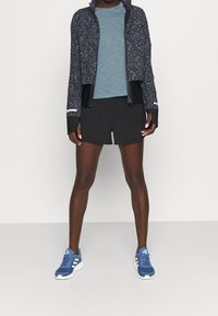 Schwarze Sportshorts, ein hellblaues, strukturiertes T-Shirt und eine dunkel gemusterte Zip-Jacke. Blaue Sportschuhe mit weißen Akzenten runden das Outfit ab.