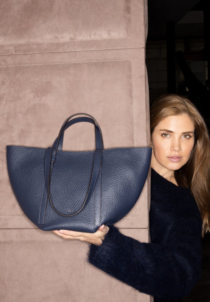 Una donna con capelli castano chiaro tiene una grande borsa navy strutturata contro uno sfondo in suede beige, indossando un maglione scuro e peloso.