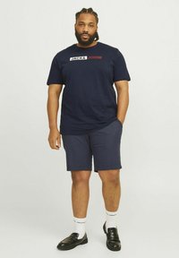 Tamsiai mėlynas medvilninis marškinėlis su baltu ir raudonu "JACK & JONES" logotipu, derinamas su tamsiomis šortais, juodais loafer'iais ir baltais kojiniais su logotipu.