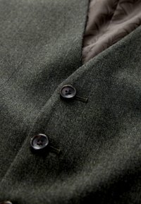 Tissu de blazer en laine vert foncé avec des boutons noirs et un aperçu de la doublure matelassée marron à l'intérieur du col.