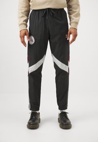 Pantalones deportivos negros con rayas diagonales en blanco y granate, cintura elástica, rayas laterales y un parche redondo en el muslo izquierdo.
