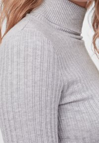 Gros plan sur un pull col roulé à côtes gris clair porté par une personne aux cheveux ondulés châtain clair.