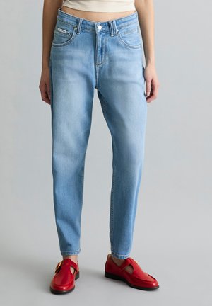 Femme portant un jean droit bleu clair et des mocassins en cuir rouge à boucle, debout sur un fond neutre.