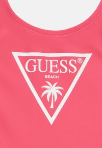 Różowa odzież kąpielowa z zaokrąglonym dekoltem, ozdobiona białym trójkątnym logo, na którym znajdują się napisy "GUESS", "BEACH" oraz grafika palmowej drzewa.