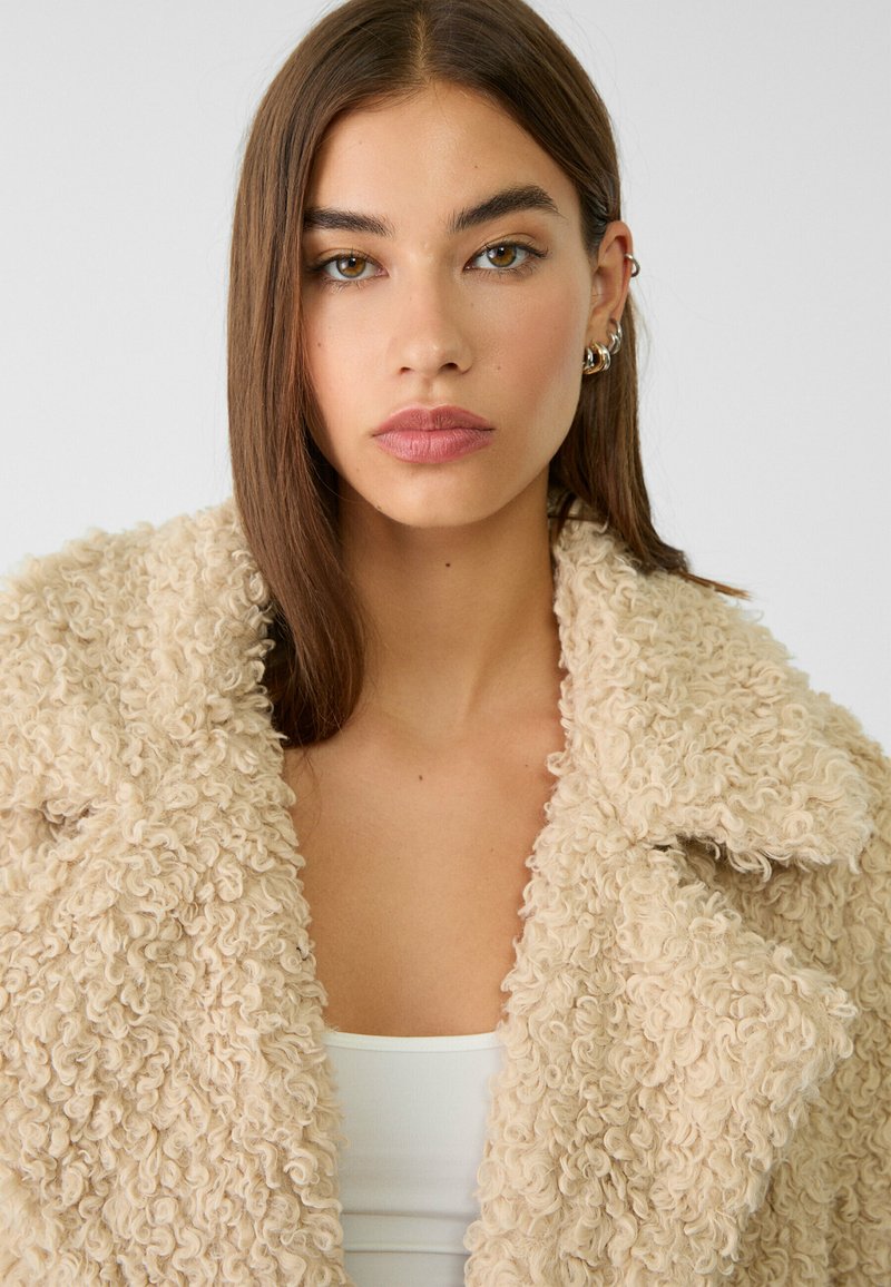 Kurtka z sztucznego futra w kolorze kremowym, o teksturowej, kręconej powierzchni. Wyróżniający się duży kołnierz i otwarty przód, w zestawieniu z białą bluzką pod spodem.