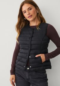 Schwarze gepolsterte Weste mit Knopfvorderseite, ausgestattet mit einem gesteppten Design und zwei Seitentaschen. Wird über einem dunkelgrauen Langarmshirt getragen.