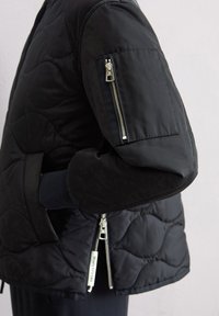 Schwarze gesteppte Pufferjacke mit einem Reißverschlusstaschen an den Ärmeln, gerippten Bündchen und sichtbaren silbernen Reißverschlussakzenten. Glatte Textur und taillierte Passform.