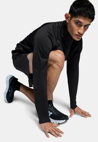 Haut de sport noir à manches longues et short, design ajusté. Chaussures de course noires avec des accents blancs. Position à genoux sur un fond clair.