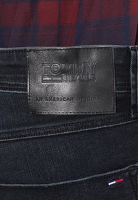 Czarna skórzana naszywka na ciemnoniebieskich dżinsach z wytłoczonym logo "TOMMY JEANS" oraz "AN AMERICAN ORIGINAL." Widoczne detale w kontrastowym szyciu.
