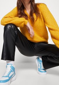 Femme portant un pull jaune texturé, un pantalon en cuir noir, des chaussettes blanches et des baskets montantes bleues et blanches, accroupie devant un fond blanc.