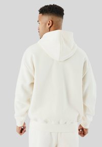 Uomo con taglio di capelli sfumati, che indossa un hoodie bianco sporco e pantaloni abbinati, in piedi di spalle rispetto alla fotocamera su uno sfondo grigio uniforme.