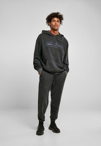 Set di felpa nera con cappuccio e pantaloni abbinati, caratterizzato da una tasca frontale a marsupio, tessuto testurizzato e logo "Urban Classics" in blu.