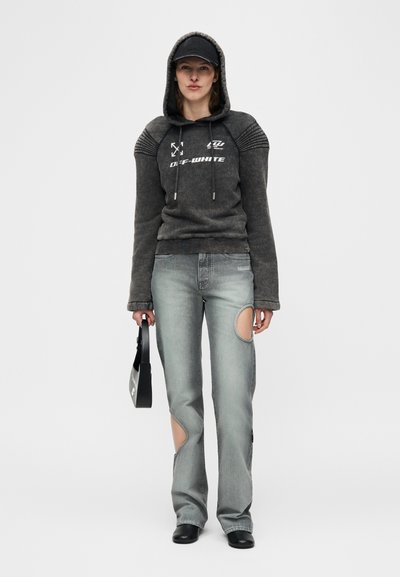 Νεαρή γυναίκα που φοράει σκουρόχρωμο φούτερ με κουκούλα με το κείμενο «OFF-WHITE», γκρι distressed τζιν με μεγάλες κοψίματα στα γόνατα, μαύρο καπέλο, μαύρα παπούτσια και κρατάει μια μαύρη τσάντα.
